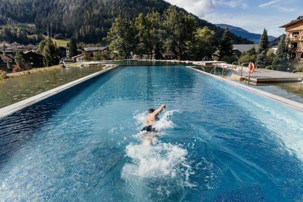 Nahaufnahme eines Mannes beim Schwimmen im Außenpool des 5-Sterne-Hotels in Bad Kleinkirchheim Ein Schwimmer zieht seine Bahnen in einem beeindruckenden Außenpool, umgeben von einer malerischen Berglandschaft. Die üppigen Bäume und sanften Hügel bieten eine ruhige Kulisse, während das klare Wasser das Licht der Sonne reflektiert.